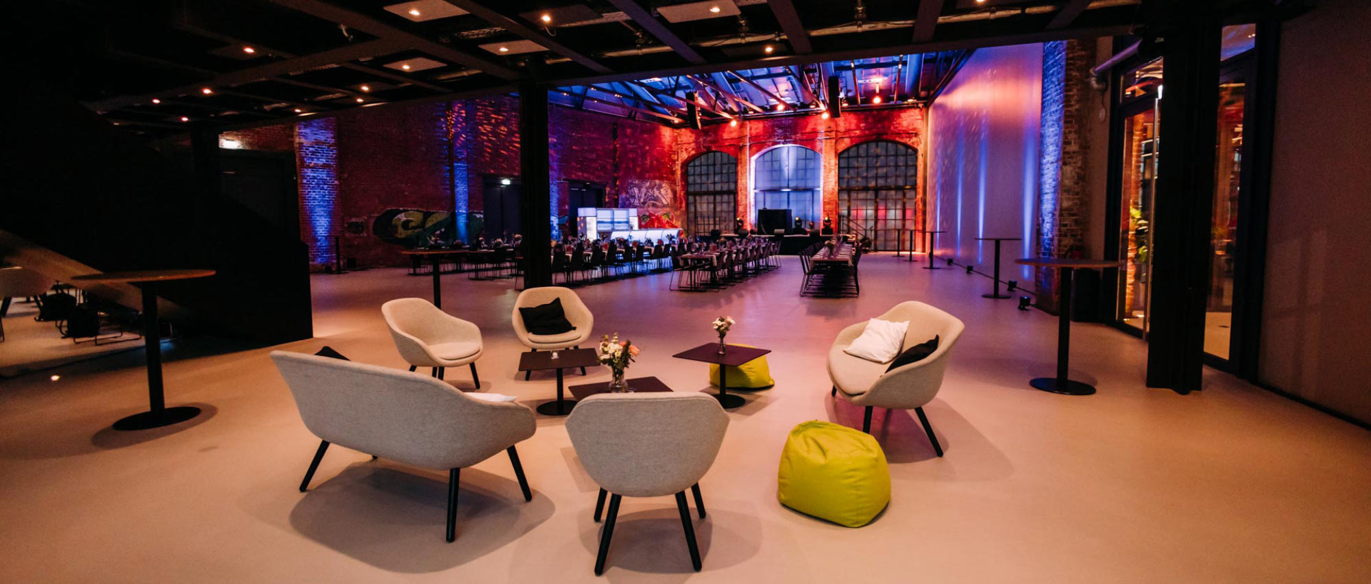 Moderner Eventsaal mit Lounge-Möbeln, Stehtischen, langer Tafel und farbiger Beleuchtung