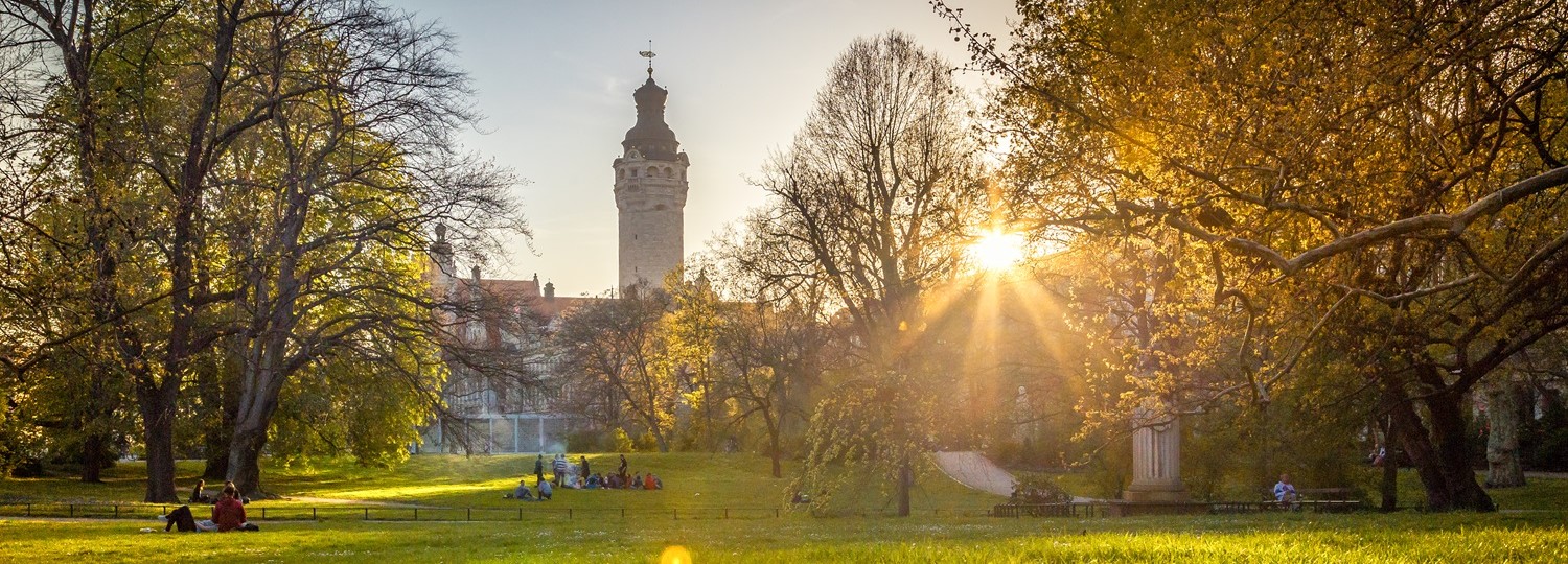 Parkwiese mit Menschen, Bäumen und Sonnenstrahlen, im Hintergrund ein historischer Turm bei Sonnenuntergang