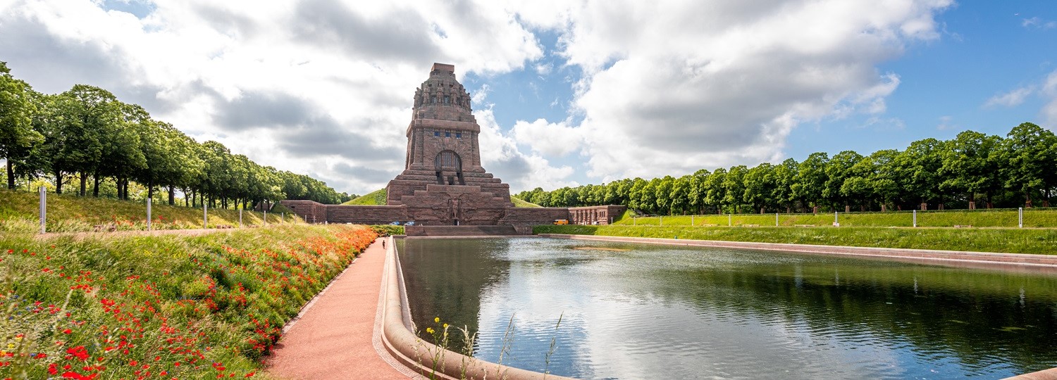 Denkmal mit Wasserbecken im Vordergrund, flankiert von Bäumen und Blumen unter bewölktem Himmel