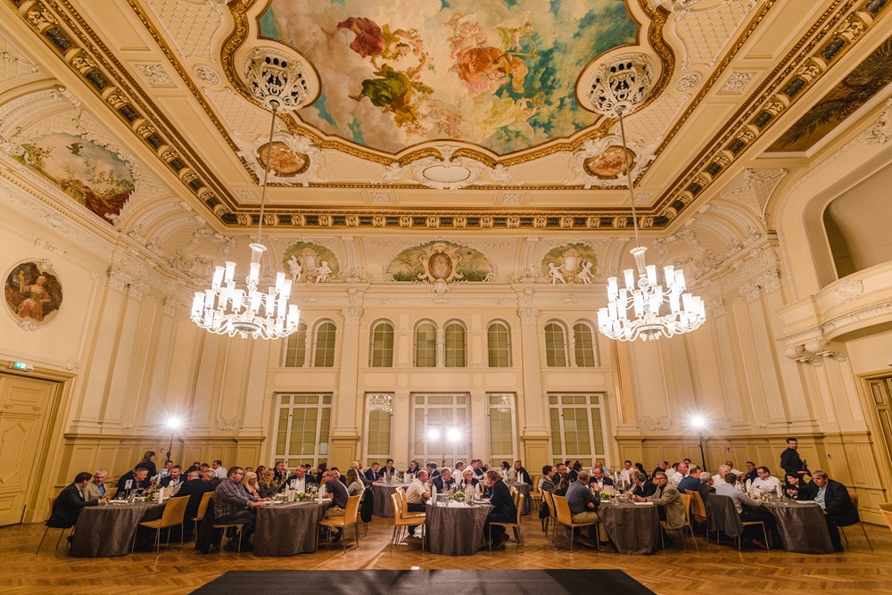 Eleganter Ballsaal mit prunkvoller Decke, Kronleuchtern und Gästen an runden Tischen bei einem Dinner