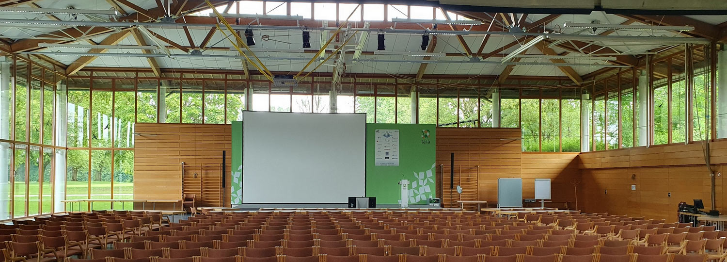 Großer, heller Konferenzraum mit vielen Stuhlreihen, Bühne, grünem Hintergrund und Projektionsfläche